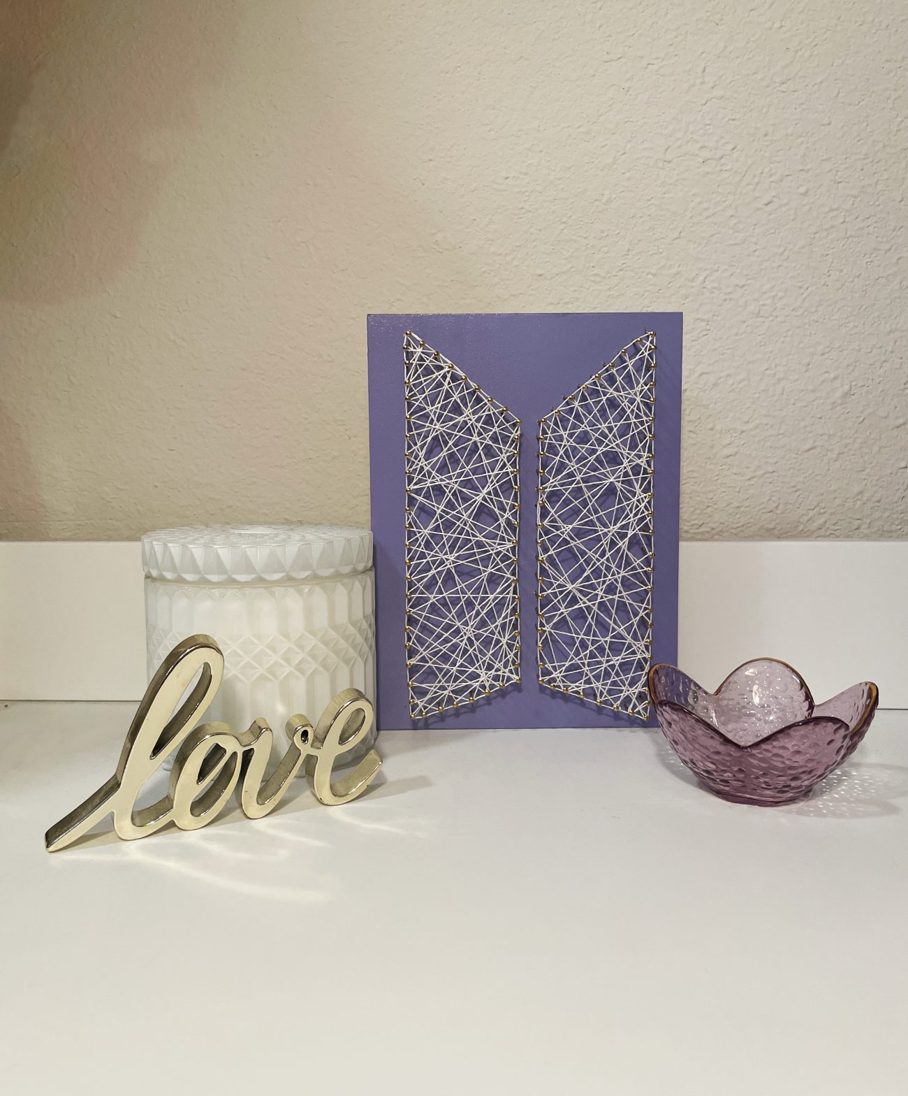 A string art rendition of BTS's modern logo done in white string on purple painted wood. It sits between a candle, a glass votive holder and a metal sign that reads 'love.'