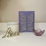 A string art rendition of BTS's modern logo done in white string on purple painted wood. It sits between a candle, a glass votive holder and a metal sign that reads 'love.'